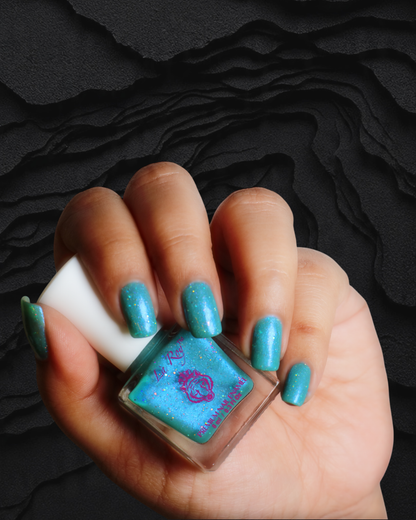 A hand holding a bottle of glittery turquoise nail polish against a dark textured background. The nails are painted with the same glittery teal polish, and the bottle label has a pink floral design.