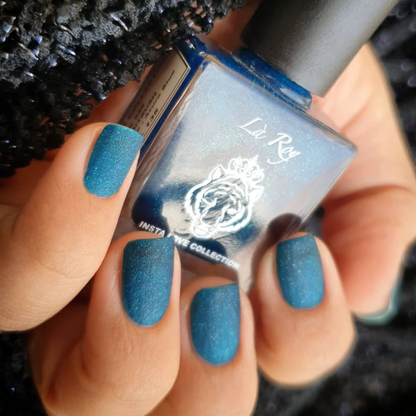 A close-up of a hand holding a bottle of nail polish, showing the nails painted with a shimmering teal blue polish. The hand is positioned in front of a black textured knit fabric, and the bottle label reads 'La Roy'.
