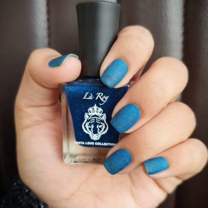 A hand holding a bottle of blue glitter nail polish, with the nails painted in a matching deep blue glitter shade. The bottle features a black cap and a blue label with the brand name 'La Roy' and 'INSTA LOVE COLLECTION' printed on it.
