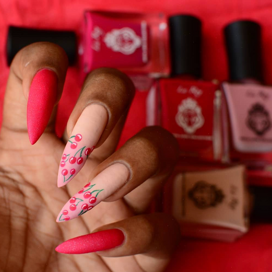 A close-up of a hand with long stiletto-shaped nails painted in bright pink and red, with cherry illustrations on two nails, posed in front of several pink nail polish bottles on a red fabric background.